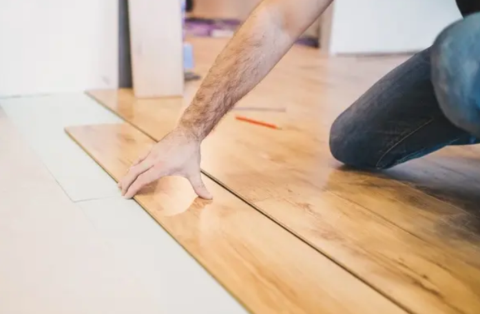 Person installing a light wood floor plank with a hand tool in a room under renovation