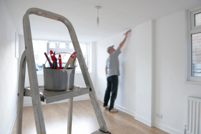 Person painting a white room beside a step ladder and bucket of paint brushes