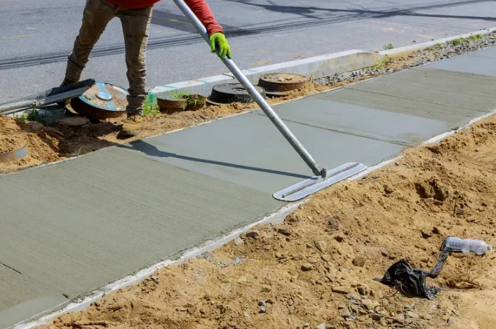 Worker smoothing wet concrete on a sidewalk with a long-handled float beside a sand pile