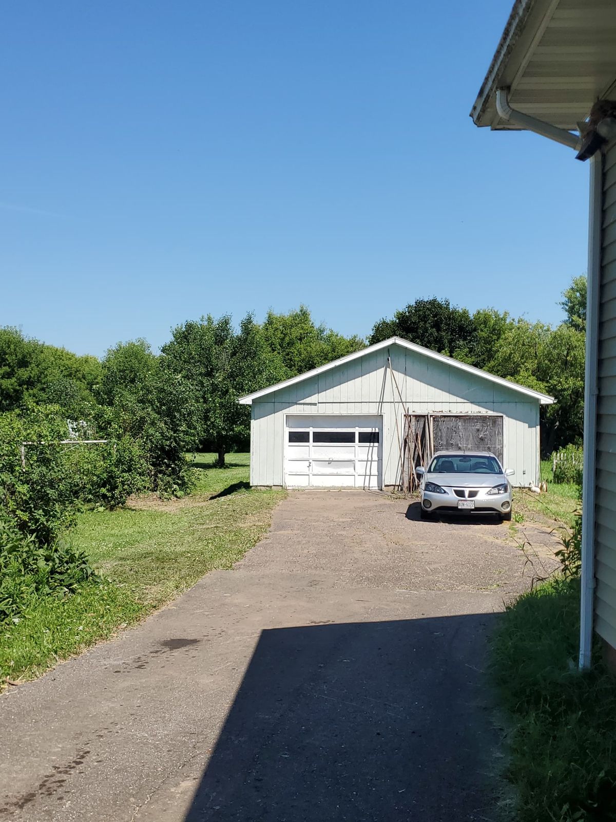 Driveway leading to a white garage with a silver car parked in front, surrounded by trees and grass.