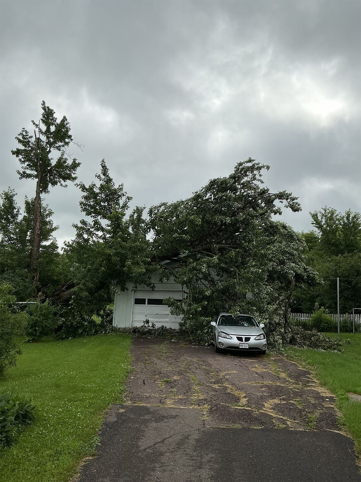 Silver car in a driveway beside a small house under cloudy skies, with trees and wet pavement.