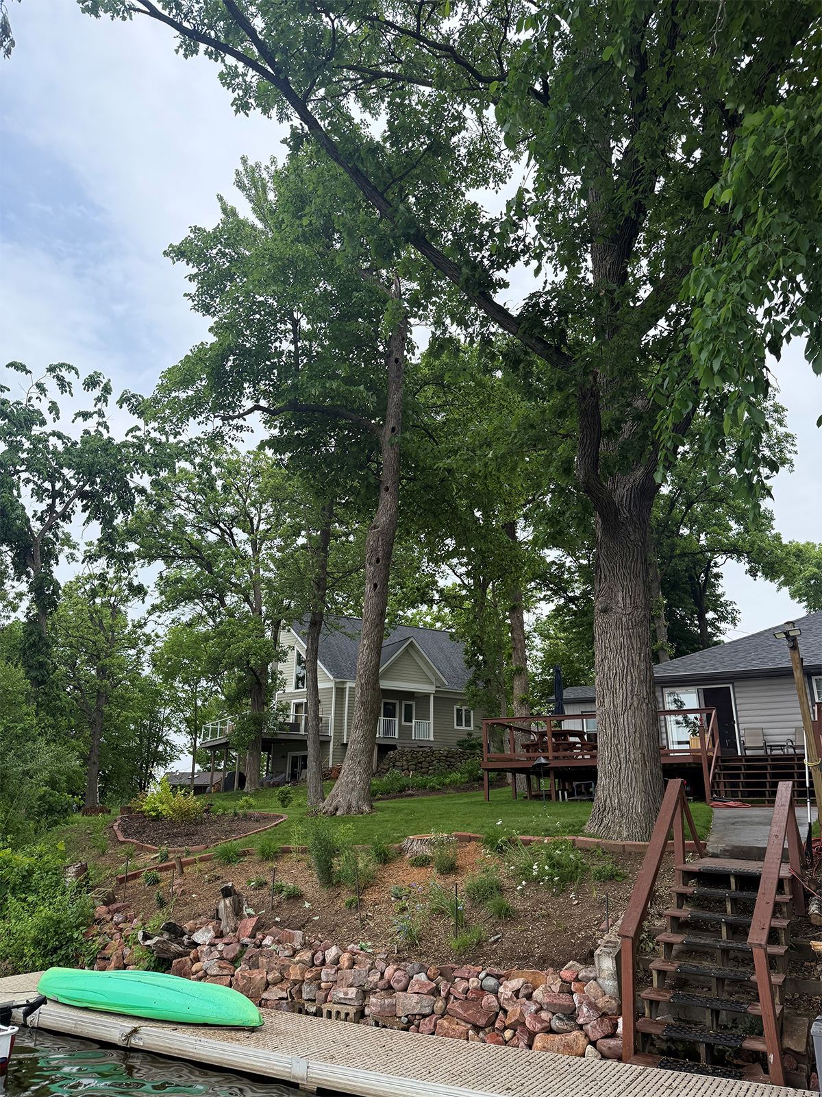 Lakeside deck and house beneath tall trees, with stone steps down to the shore and a green kayak nearby