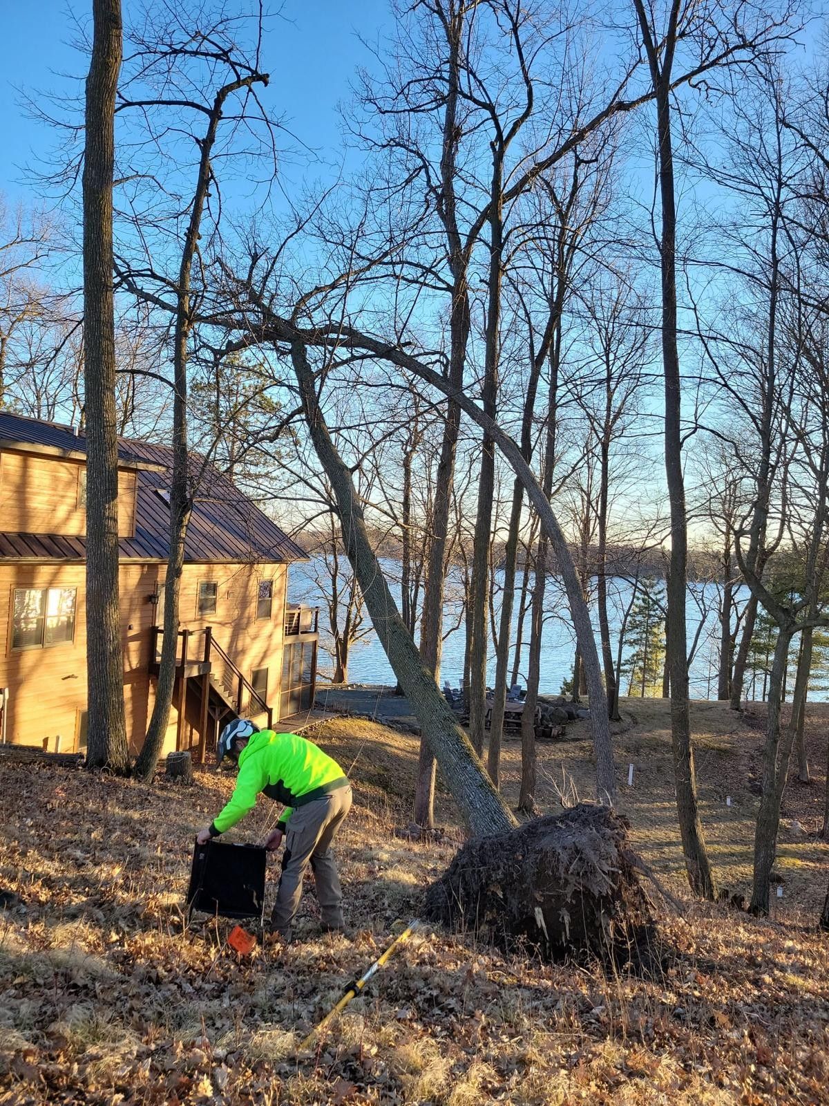 Person in a green jacket tending a black dog beside a lakeside cabin in a wooded yard.