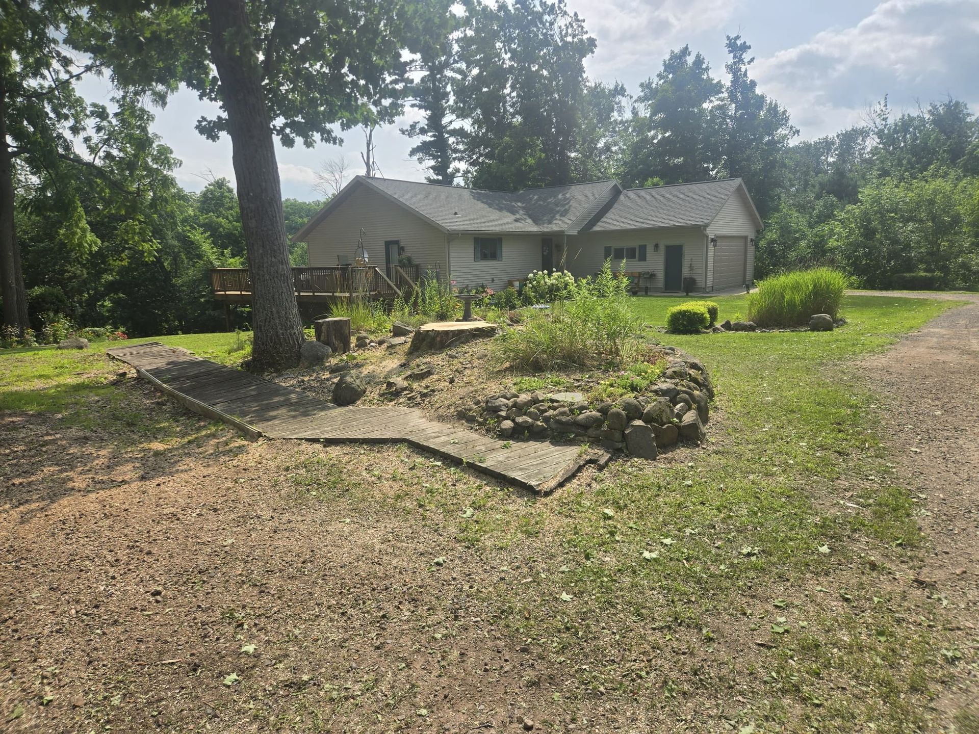 Rural house with a stone garden bed, large tree, and grassy driveway under bright sunlight