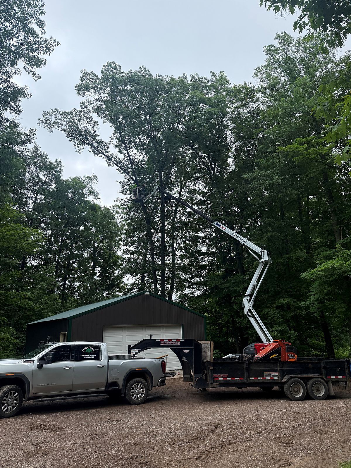 Two trucks and a bucket lift truck parked beside a small shed in a wooded yard
