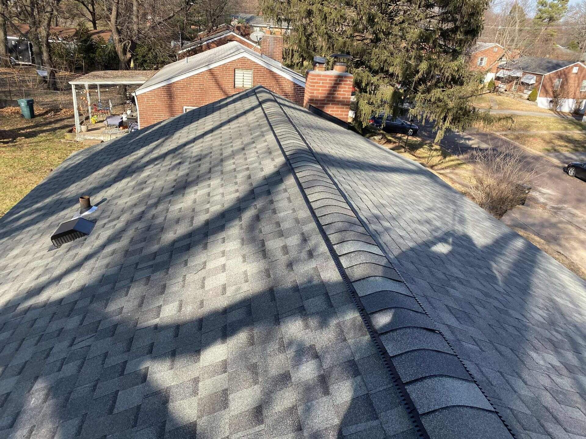 View of a gray shingled roof with a central ridge. Surrounding trees and other houses are visible.