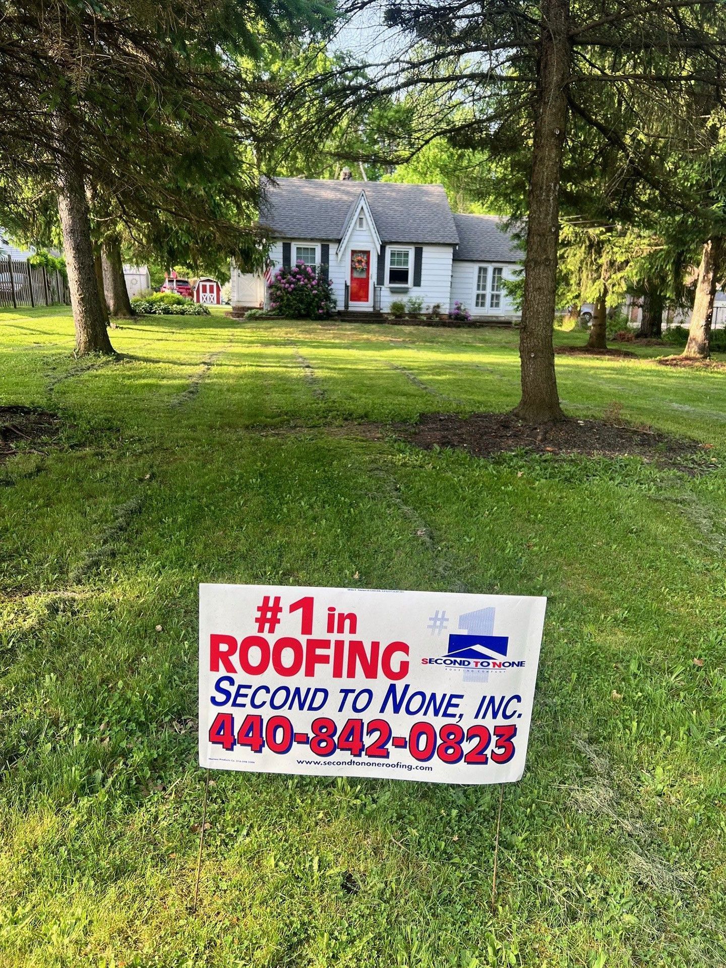 A roofing sign is sitting in the grass in front of a house.