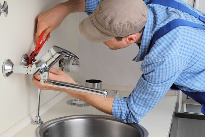 Plumber installing a faucet over a stainless steel sink, using pliers.