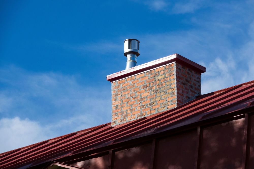 Brick chimney with metal cap on a red roof against a blue sky.