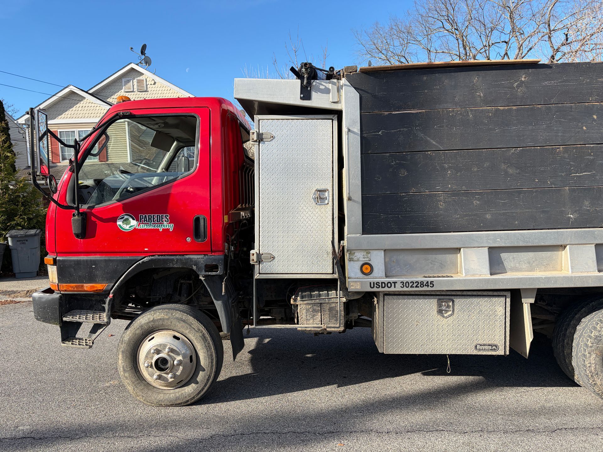 Red dump truck parked on a street; silver storage compartments, black bed.