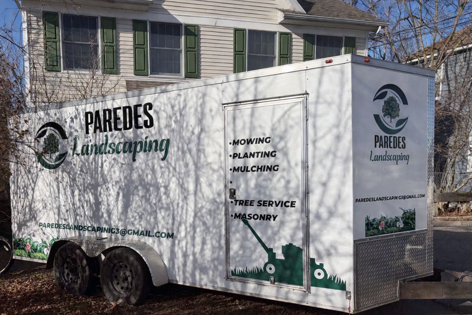 White trailer for Paredes Landscaping with logo, services, and contact info, parked in front of a house.