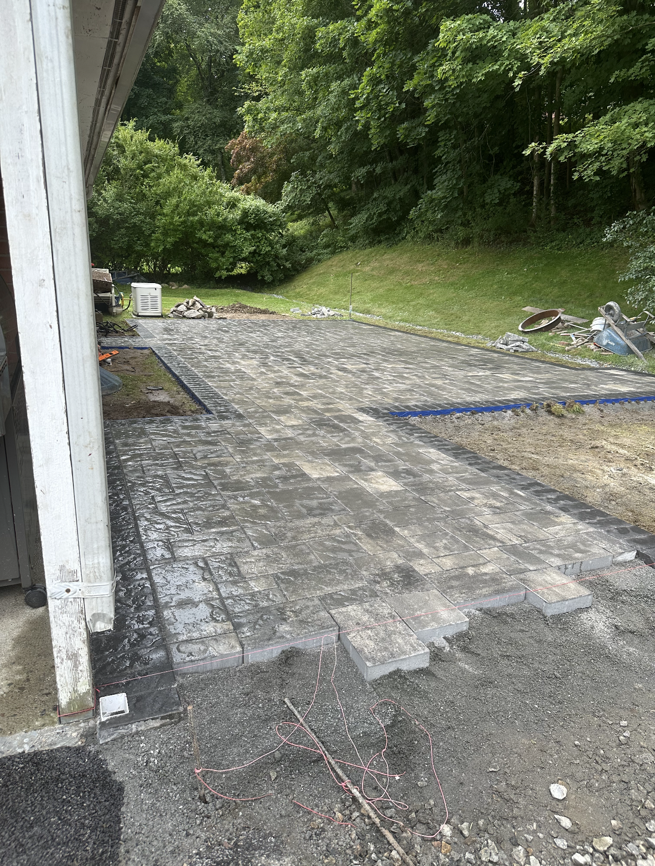 Patio construction in progress. Gray pavers laid out, gravel base, building and trees in background.