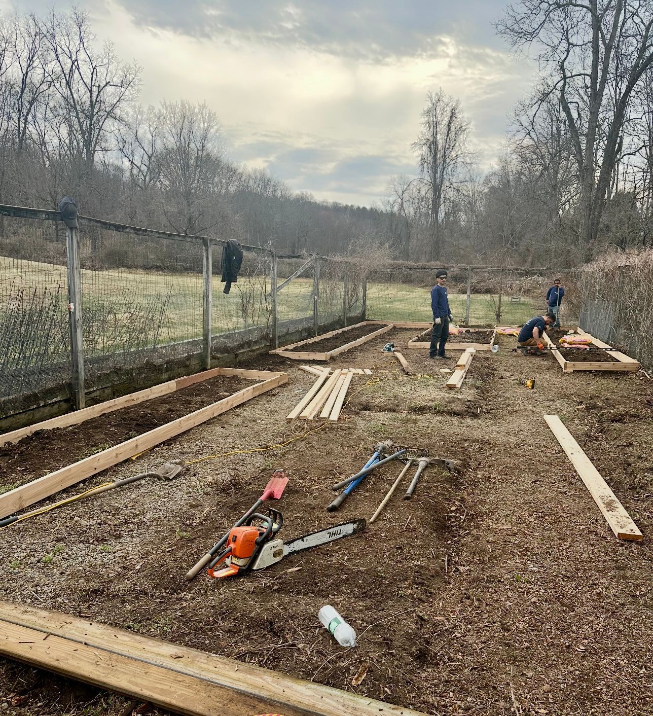 People building raised garden beds outdoors with tools and wood. Cloudy sky.