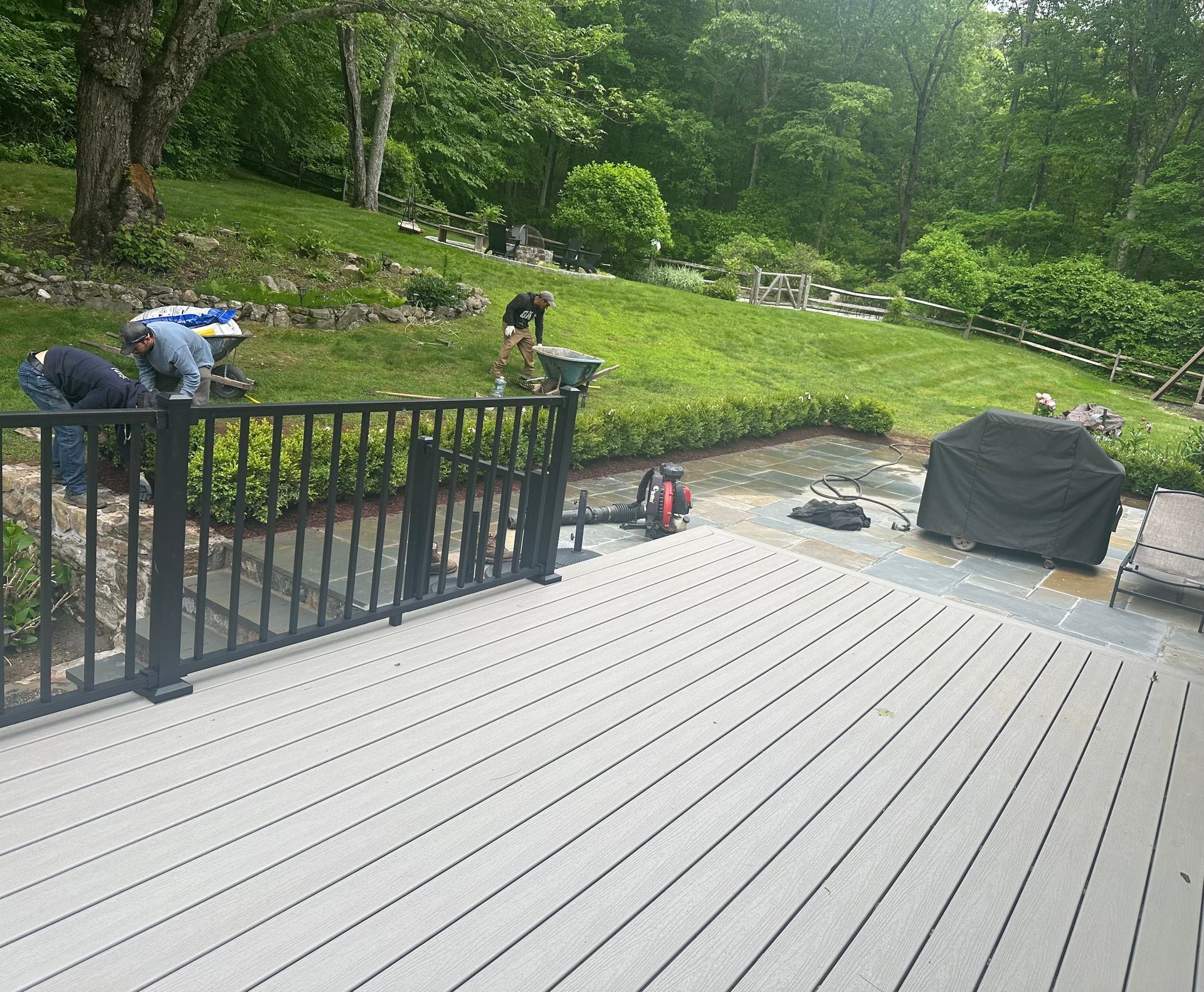 Workers installing a black railing on a gray deck overlooking a yard and woods.
