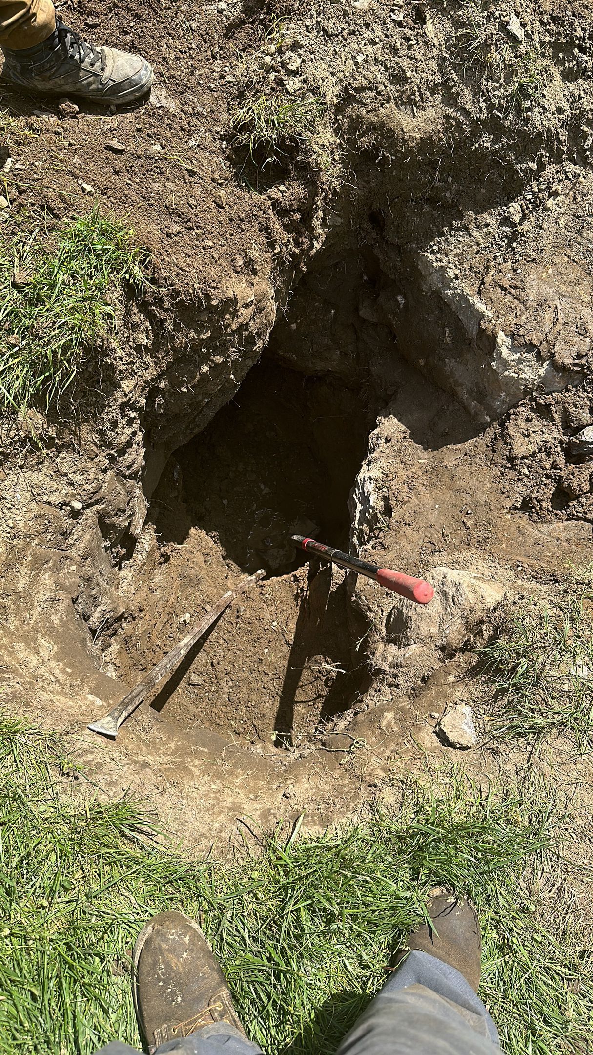 Hole dug in dirt with a red-handled tool inside. Feet with dirty boots visible in the foreground.
