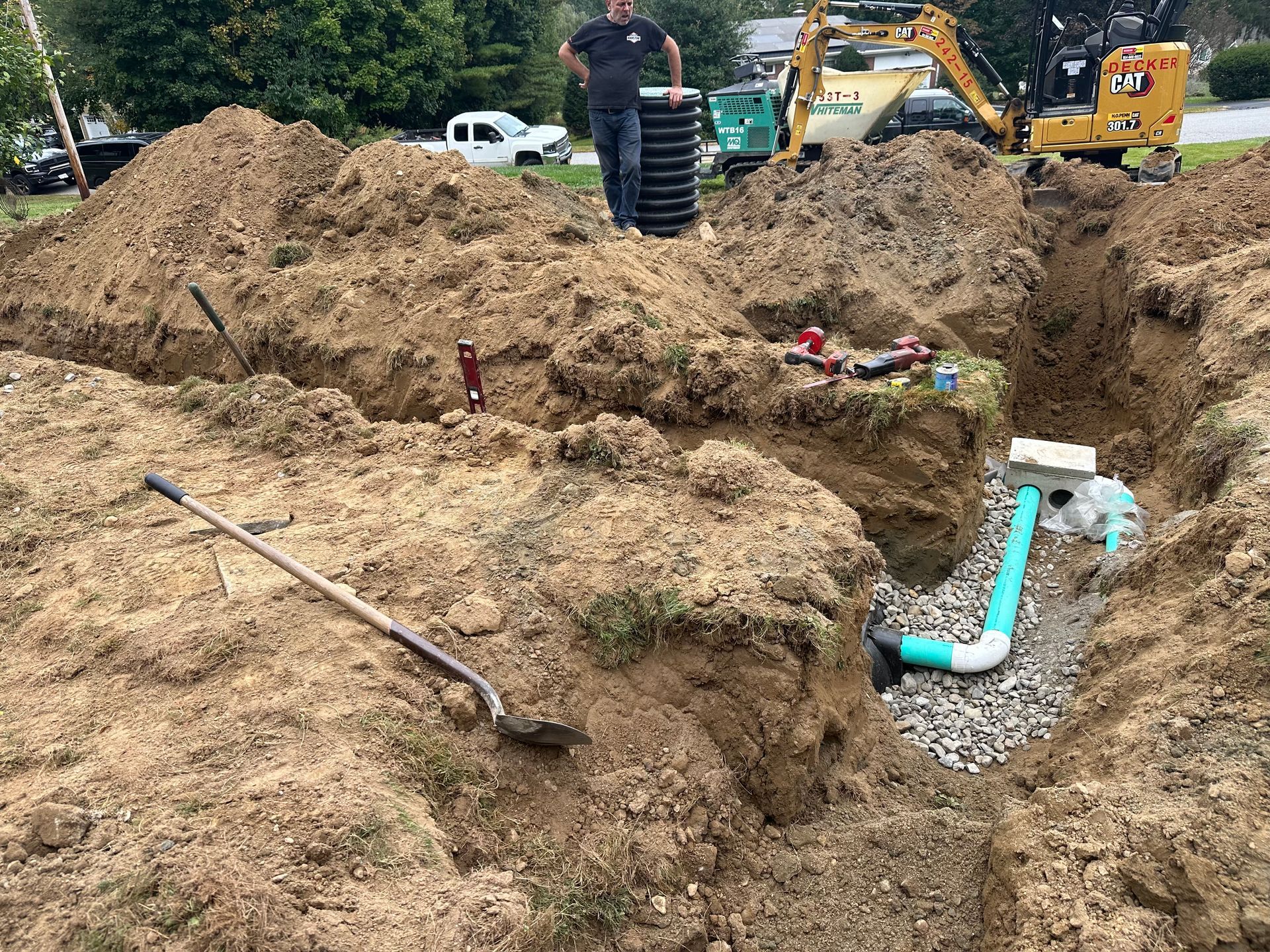 Construction site: man near excavated trenches, plumbing visible, equipment, and piles of dirt.