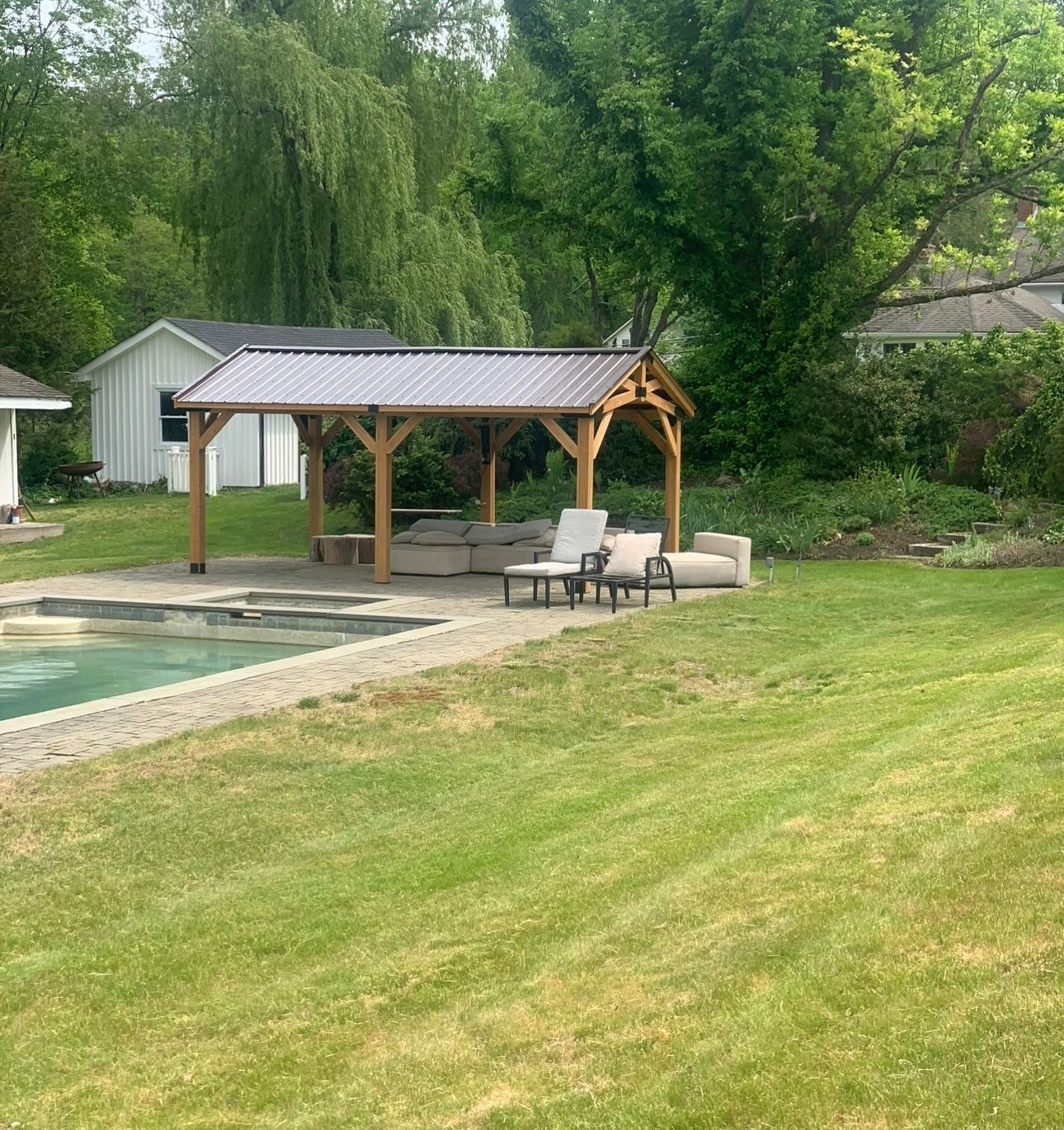 Gazebo with seating next to a pool and lawn, with trees and a white building in the background.