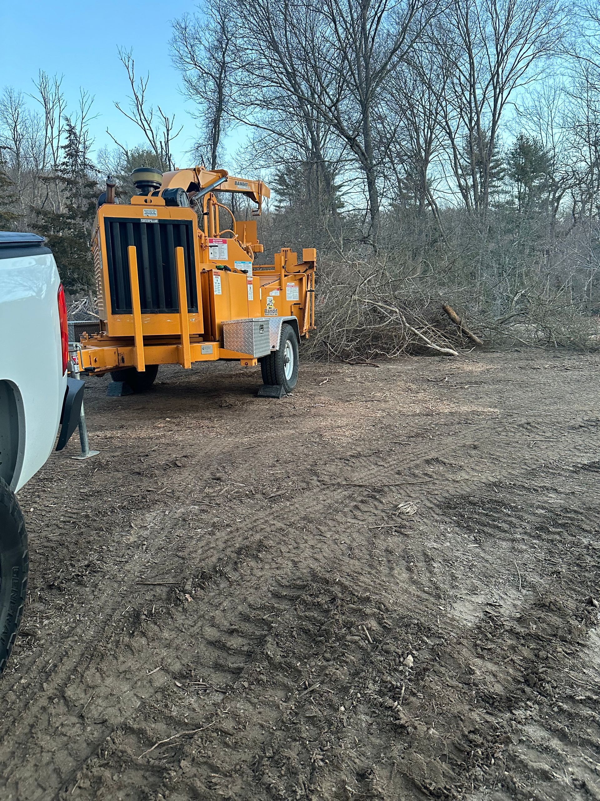 Orange wood chipper next to a pile of branches on a brown, muddy ground.