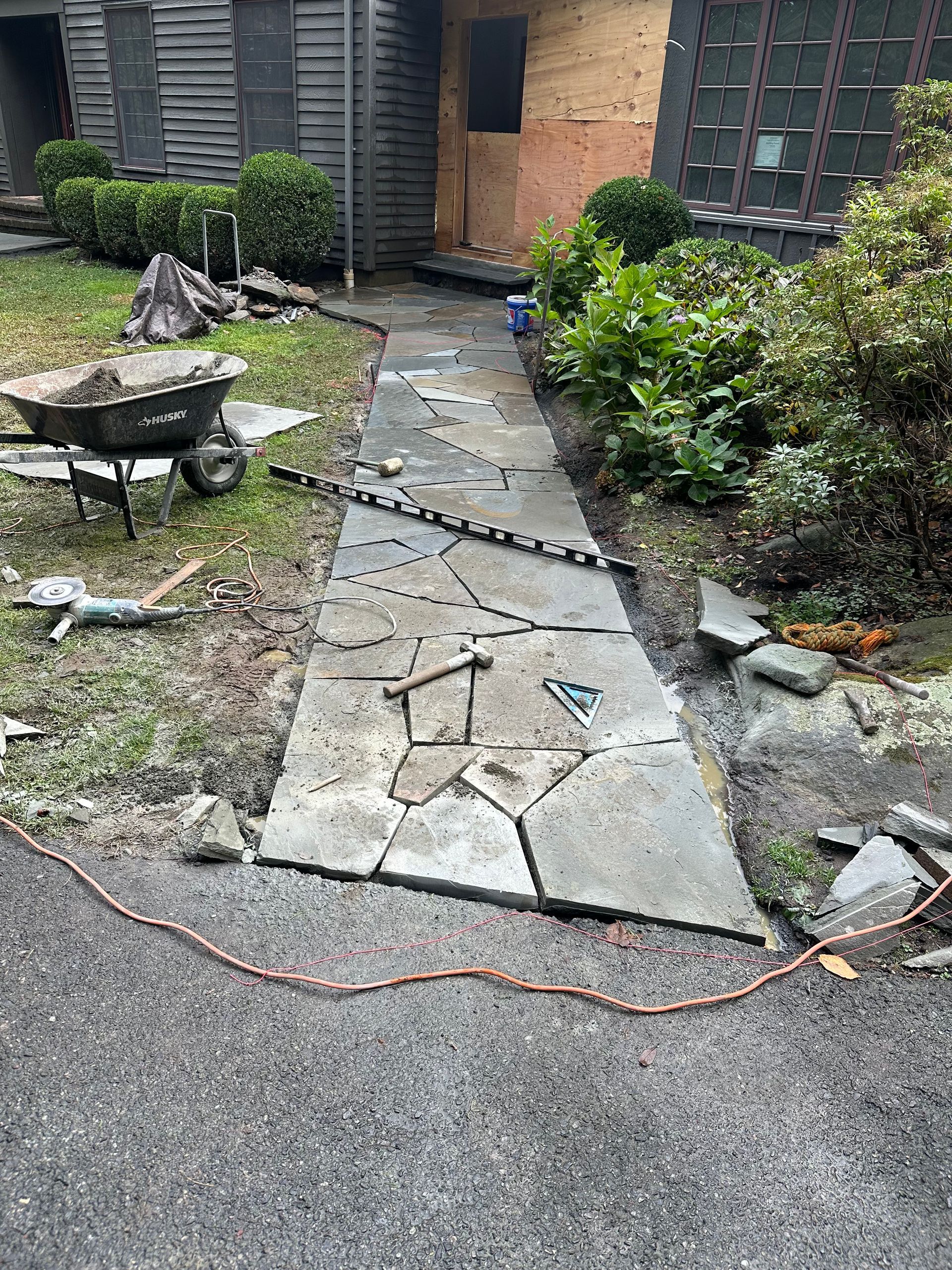 Path with broken stone slabs under construction, surrounded by tools and greenery.