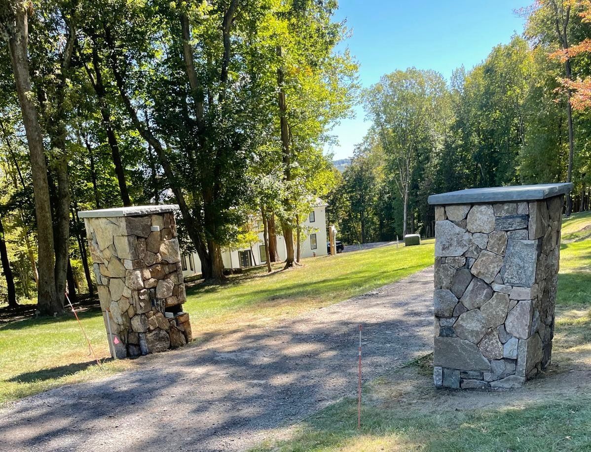 Stone pillars frame a grassy path leading to a white building, surrounded by trees.