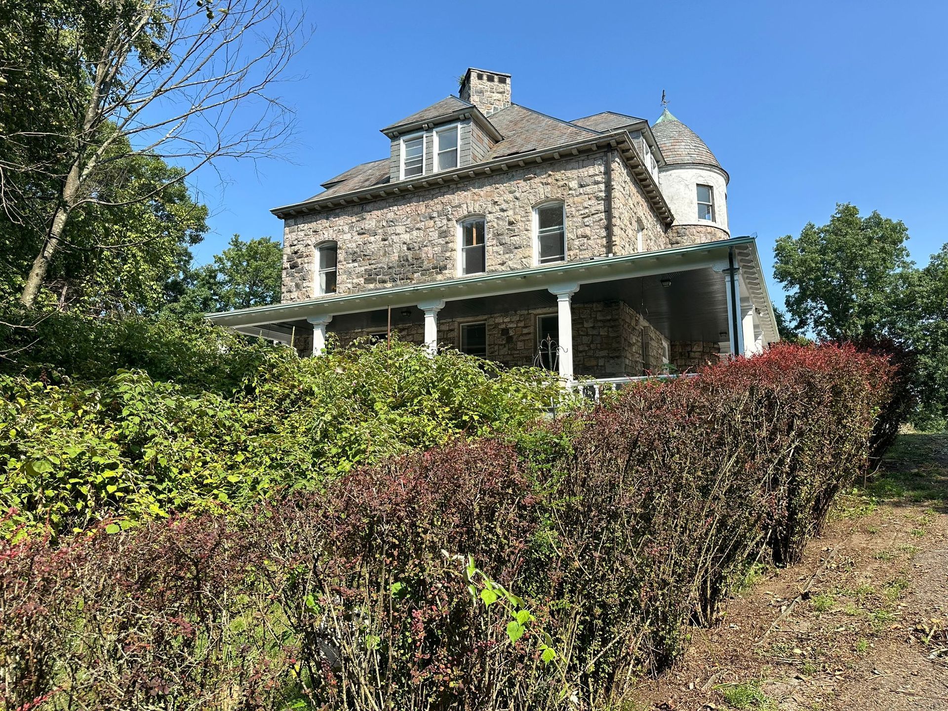 Stone house with porch, small tower, and overgrown shrubs on a sunny day.
