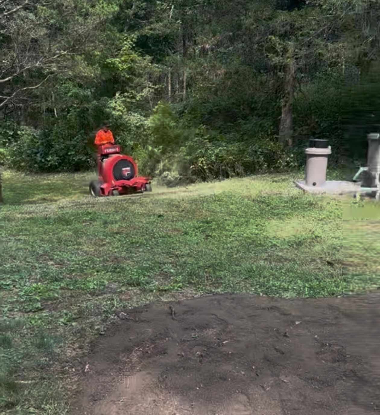 Person operating a red blower on a grassy area near trees.