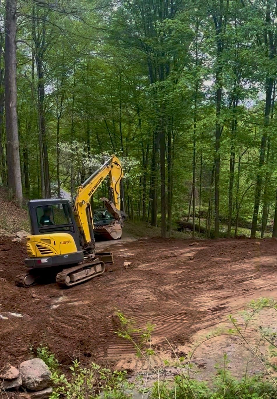 Yellow excavator on a dirt clearing in a wooded area, trees in background.