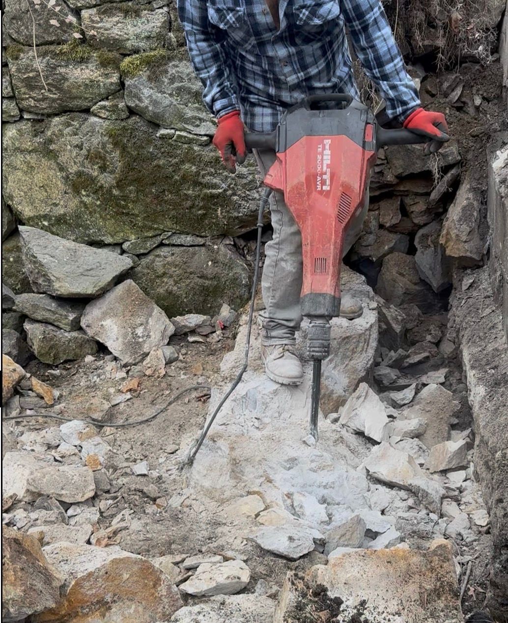 Person using a jackhammer to break up a concrete block in an outdoor setting.