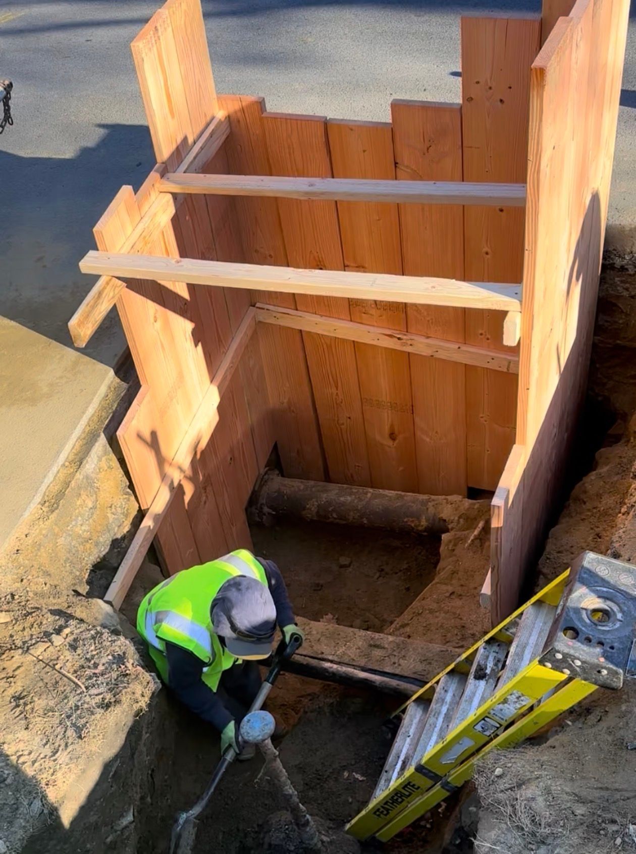 Construction worker in a trench with wooden support structure. Yellow vest, ladder, shovel visible.