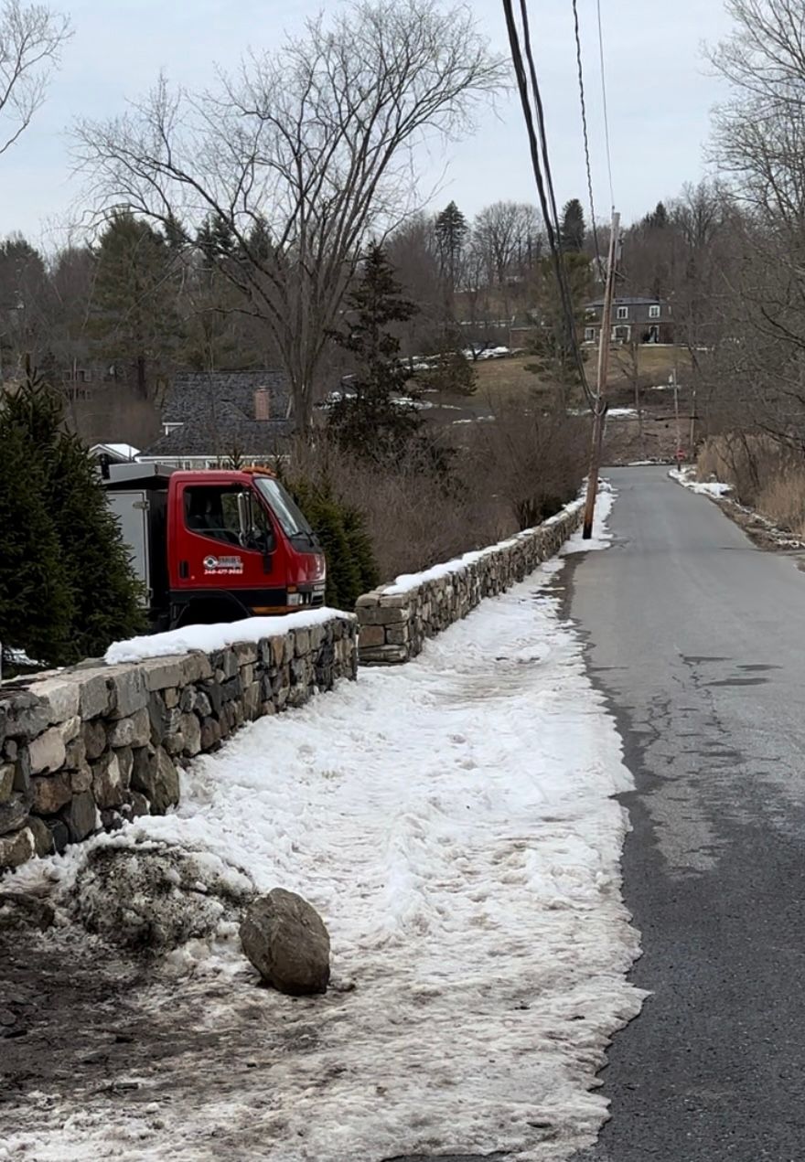 Red truck parked near a stone wall with snow, on a narrow road in a residential area.