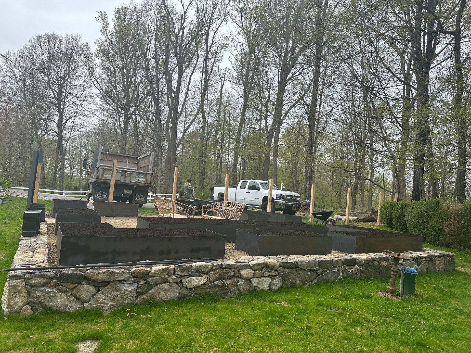 Raised garden beds with stone border; a truck and equipment are nearby; trees in the background.