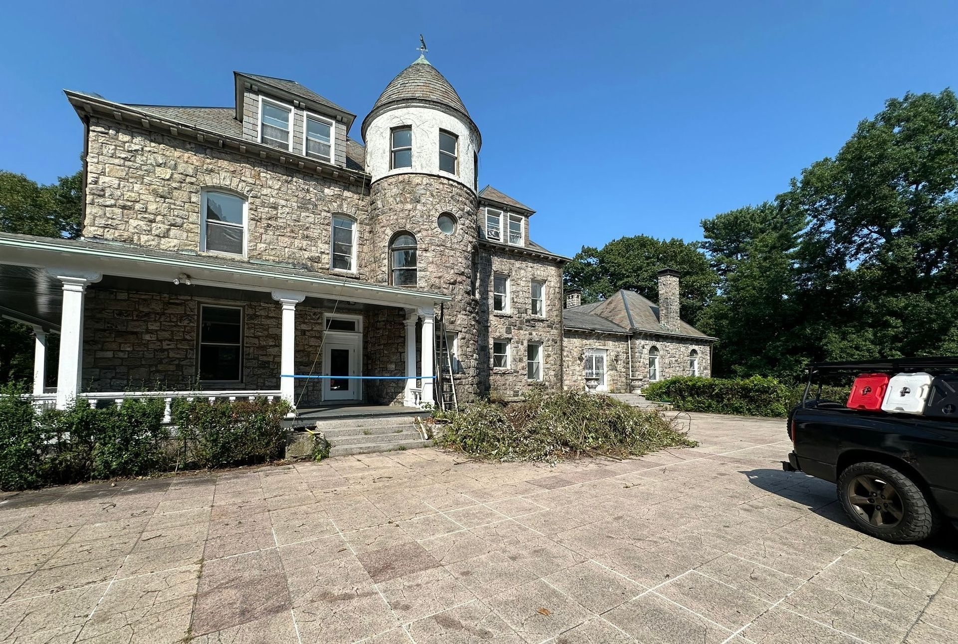 Stone mansion with a tower, a porch, and a black car in the driveway on a sunny day.