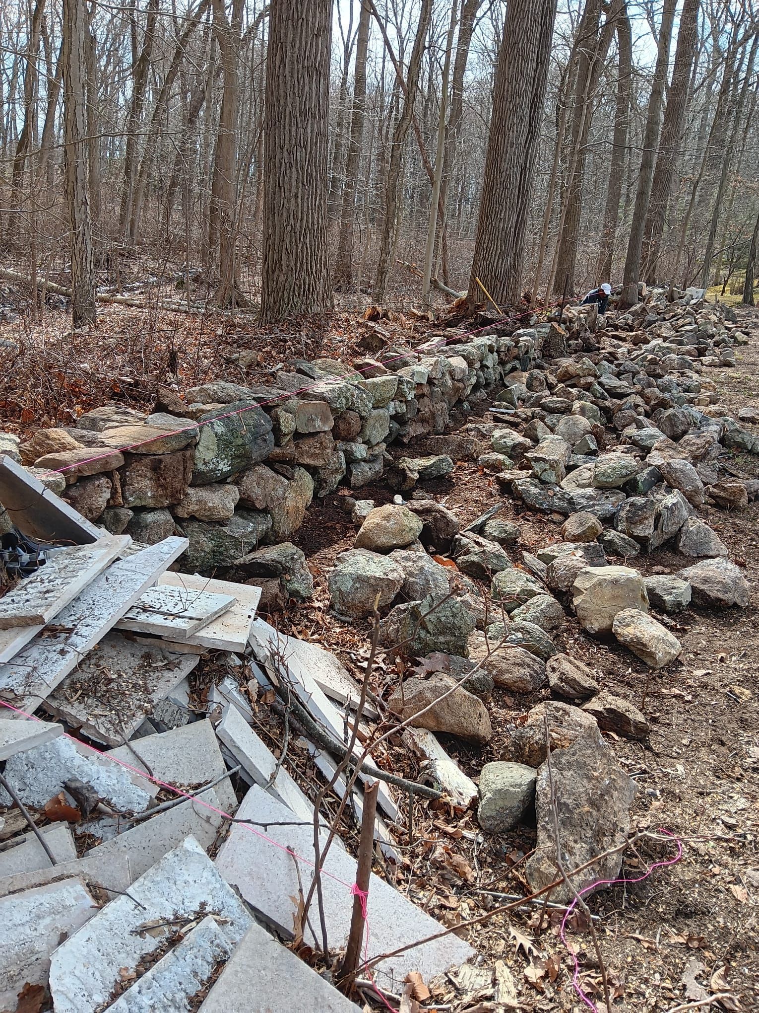 Stone wall in a forest setting with a pile of broken tiles in the foreground.