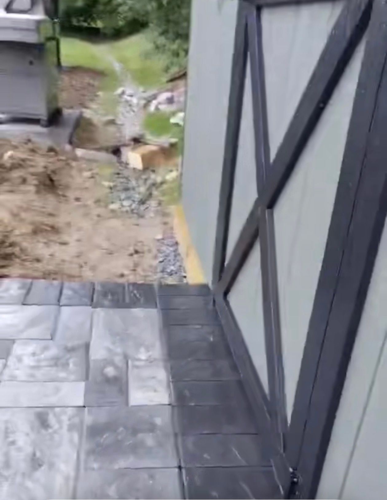 Paver walkway leading to door with X-shaped black frame; dirt and grass in the background.