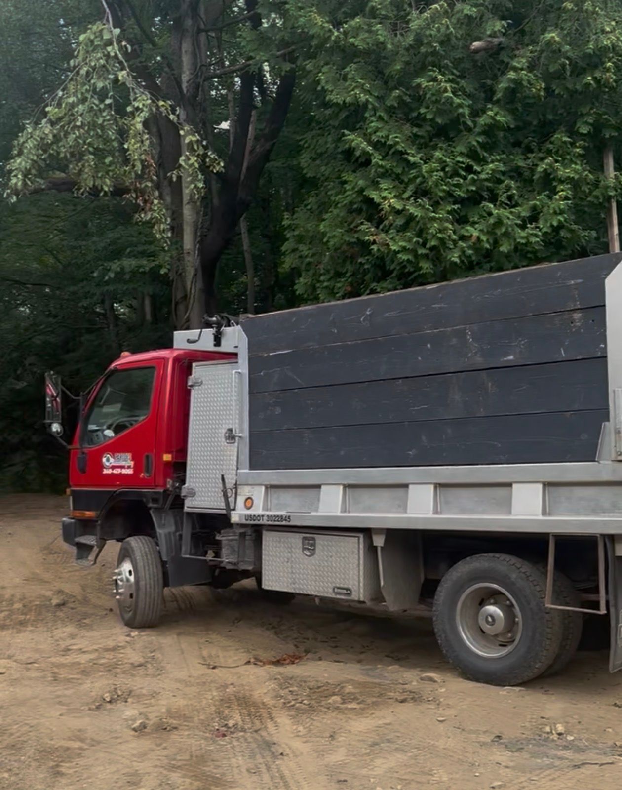 Red dump truck parked on a dirt road, trees in the background.