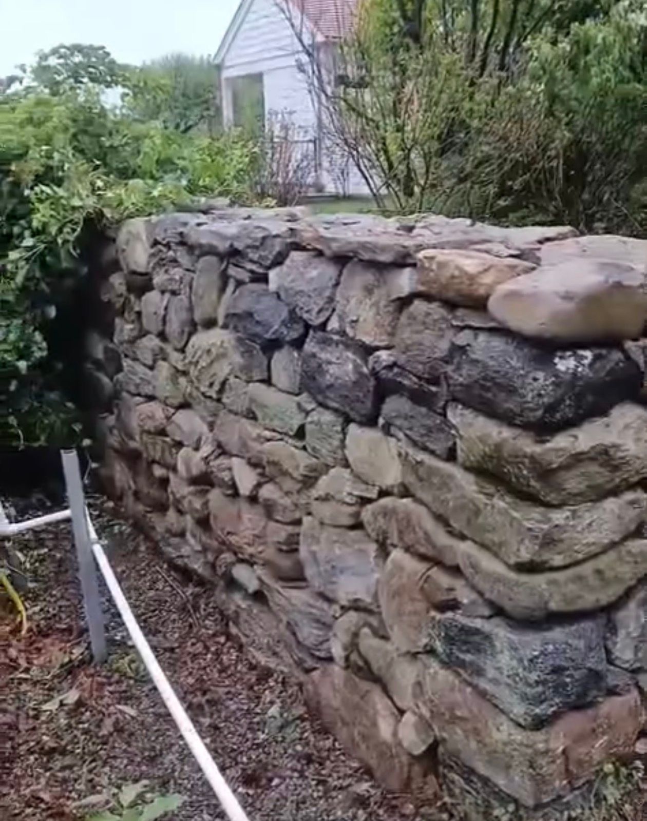 Stone wall in garden, light and dark gray stones, with white house visible in background.