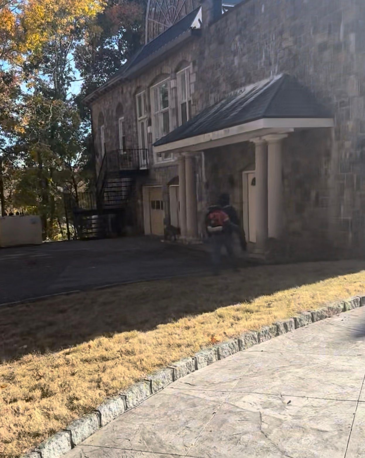 Person using a leaf blower in front of a stone building with a small awning; autumn setting.