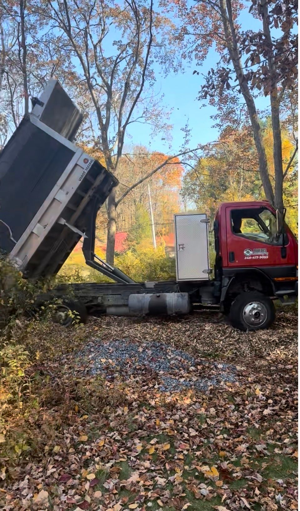 Red dump truck with raised bed, unloading debris in a wooded area with autumn leaves.