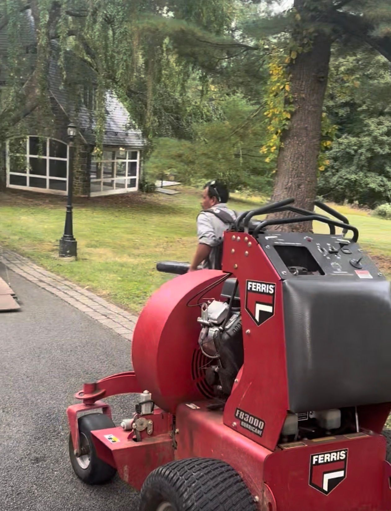 A person on a red Ferris lawn mower on a paved path near a small building with large windows.
