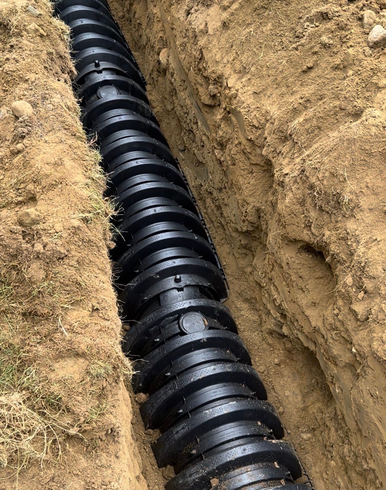 Black corrugated drainage pipe in a narrow trench dug in brown soil.