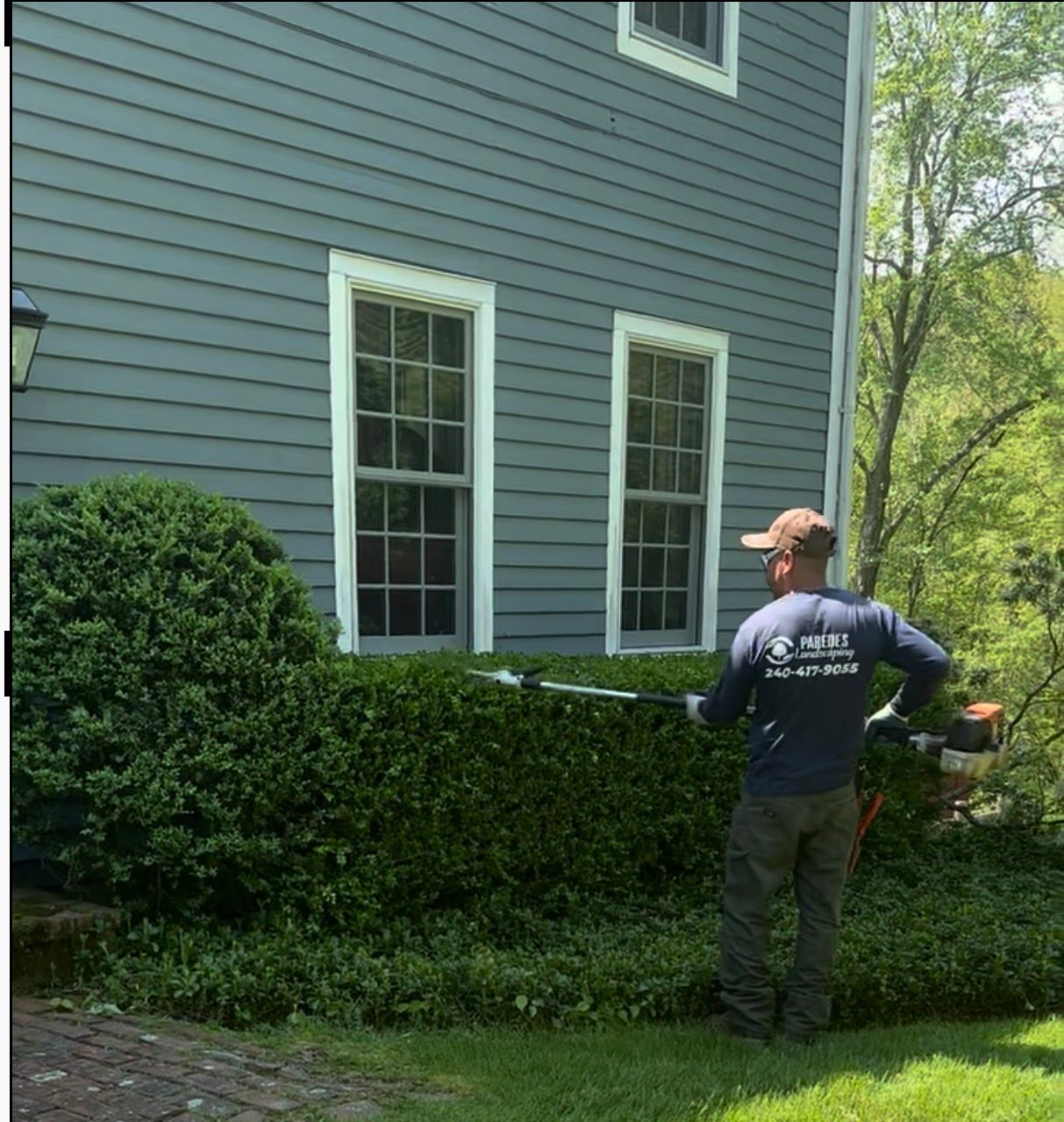 Man trimming a hedge with a power tool near a house with gray siding and windows.