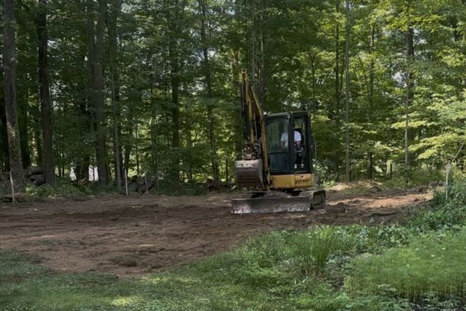 Mini excavator clearing land near a forest, brown soil, green grass, trees in background.