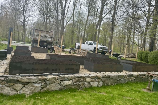 Raised garden beds filled with dark soil, stone wall in front, with trucks and trees in the background.