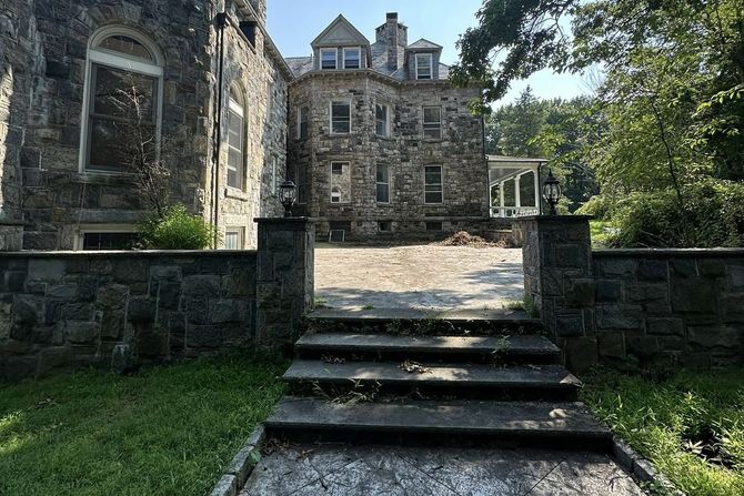 Stone mansion, front view, with steps and stone wall leading to the entrance. Overcast sky.