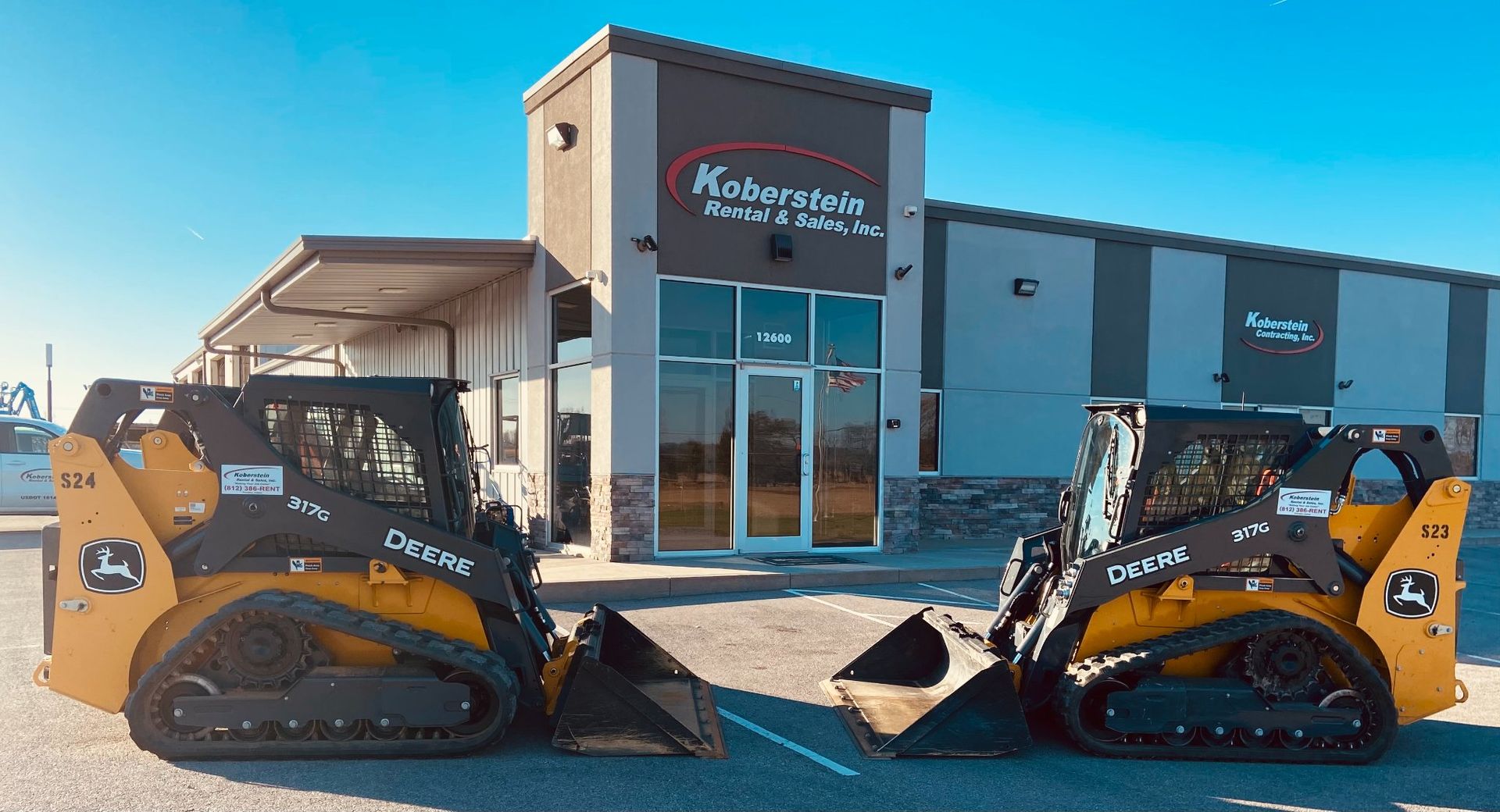 Two John Deere skid steer loaders parked in front of a Kobayakawa store on a sunny day.