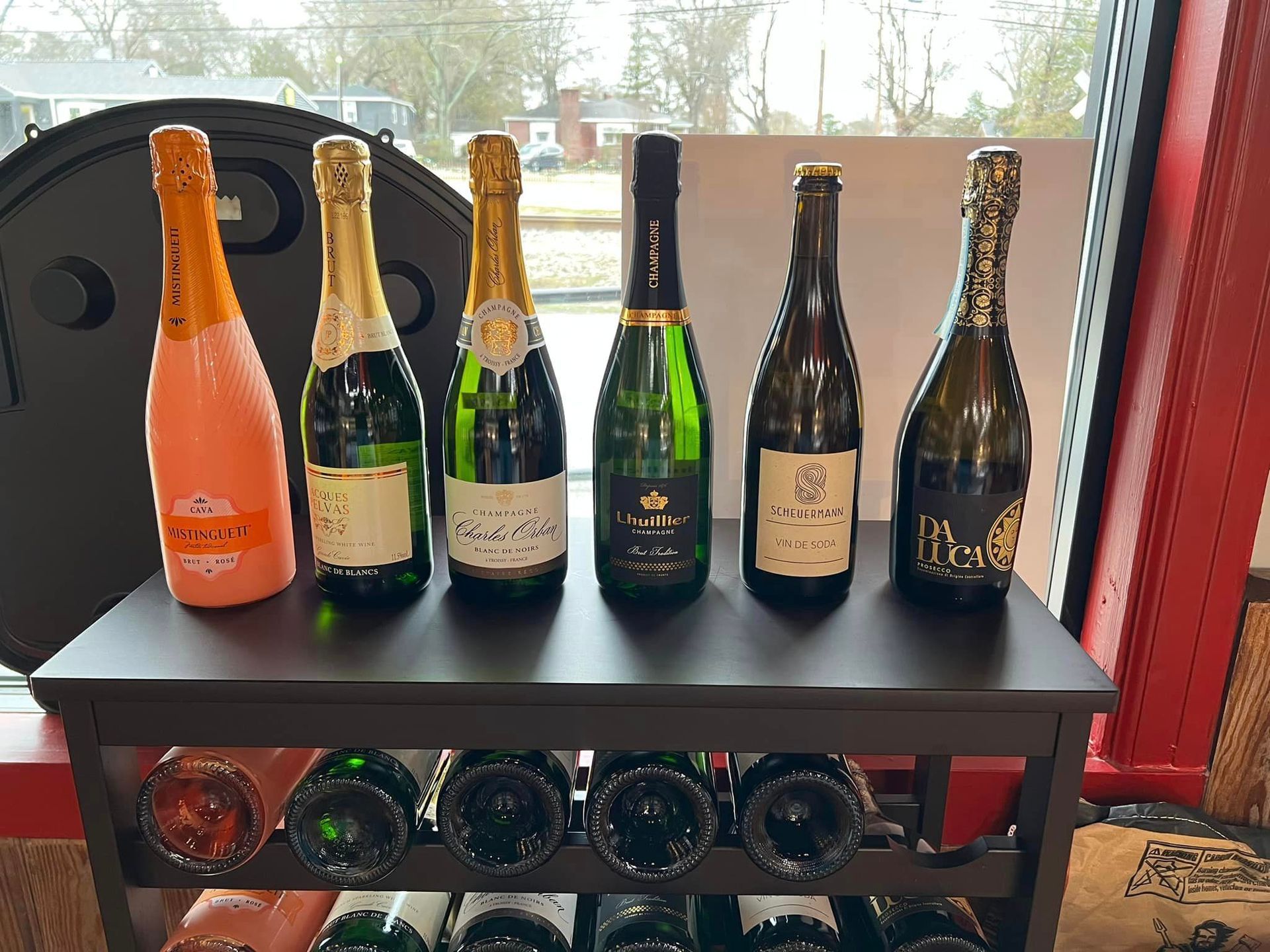 Bottles of champagne on a shelf, displayed near a window. Some bottles are pink or gold, and green.