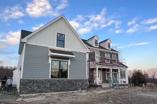 A two-story house under construction with gray siding, stone base, and a covered porch against a blue sky