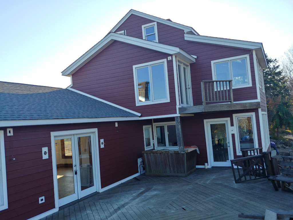 A multi-story maroon home with white trim, featuring a deck, large windows, and a paved patio area.