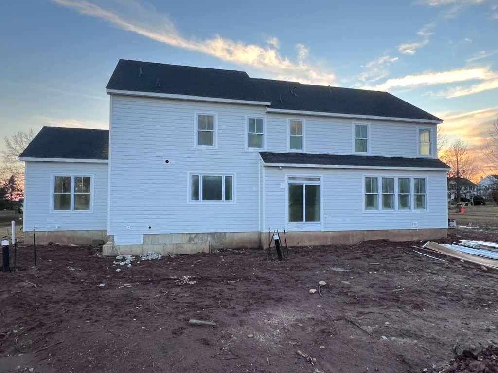 A newly constructed, two-story light blue house with a dark shingle roof stands on an unfinished dirt yard at dusk.