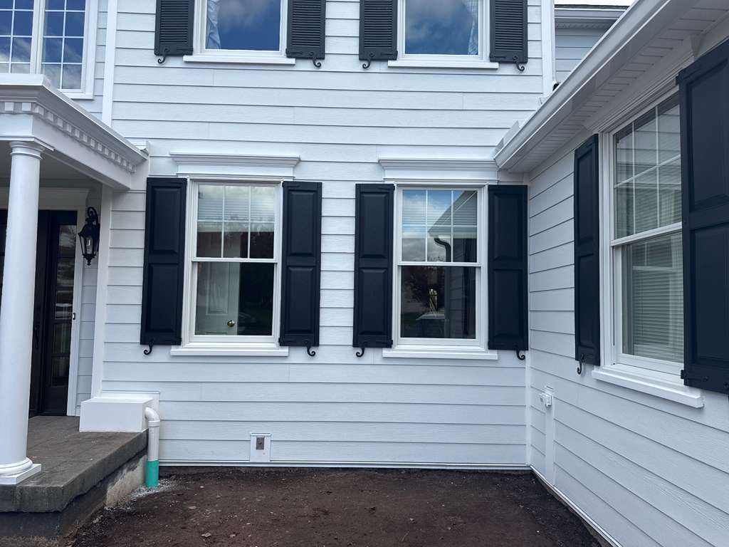 A white house exterior featuring two windows with black shutters, a porch column, and a dirt yard in the foreground.